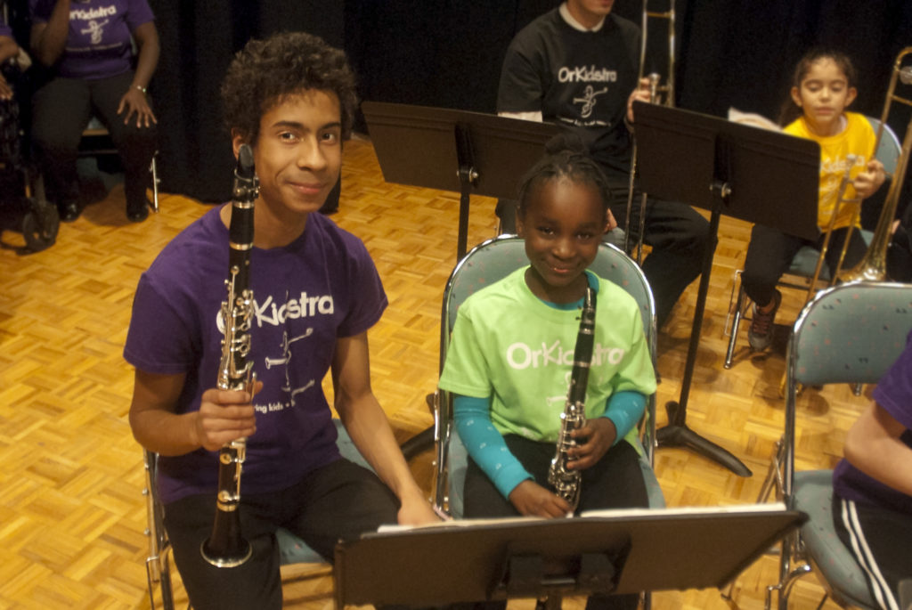 A photo of a teenage-boy and a younger girl smiling at the camera. They are both holding clarinets.
