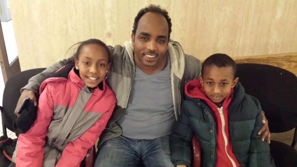 A photo of a family consisting of a father and two younger children smiling at the camera. The father is named Melake and two children are Shan and Sham.